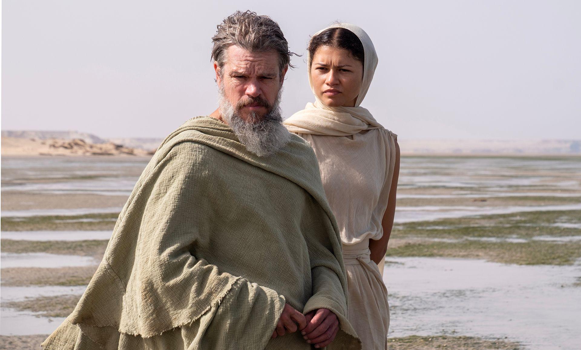 Matt Damon and Zendaya in the Odyssey both look towards camera while wearing ancient Greece-style robes against a stark marshy background and a grey sky.