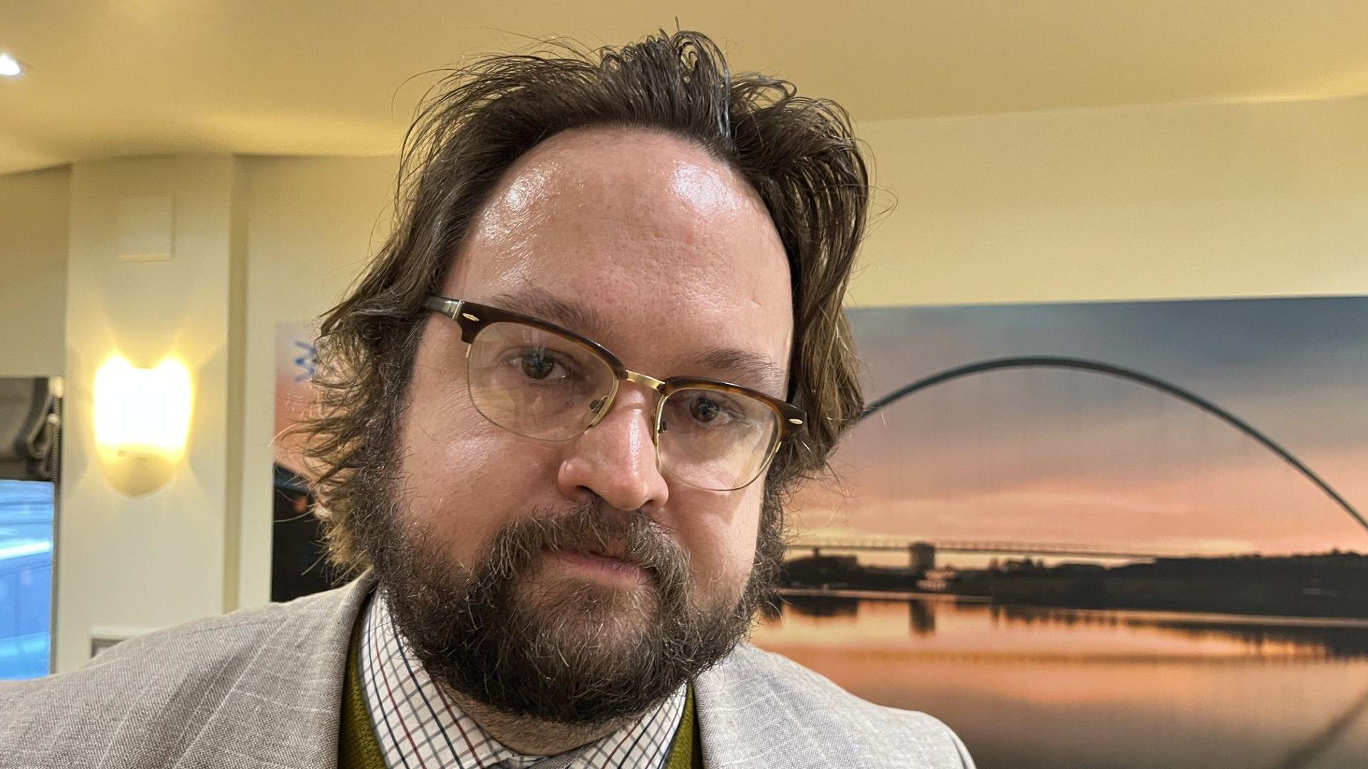 Matt Storey has messy brown hair and a beard, is wearing glasses and stands to the left of the frame. He's wearing a light suit jacket and shirt with a check pattern. Behind him is a canvas of an arched bridge over some water at sunset