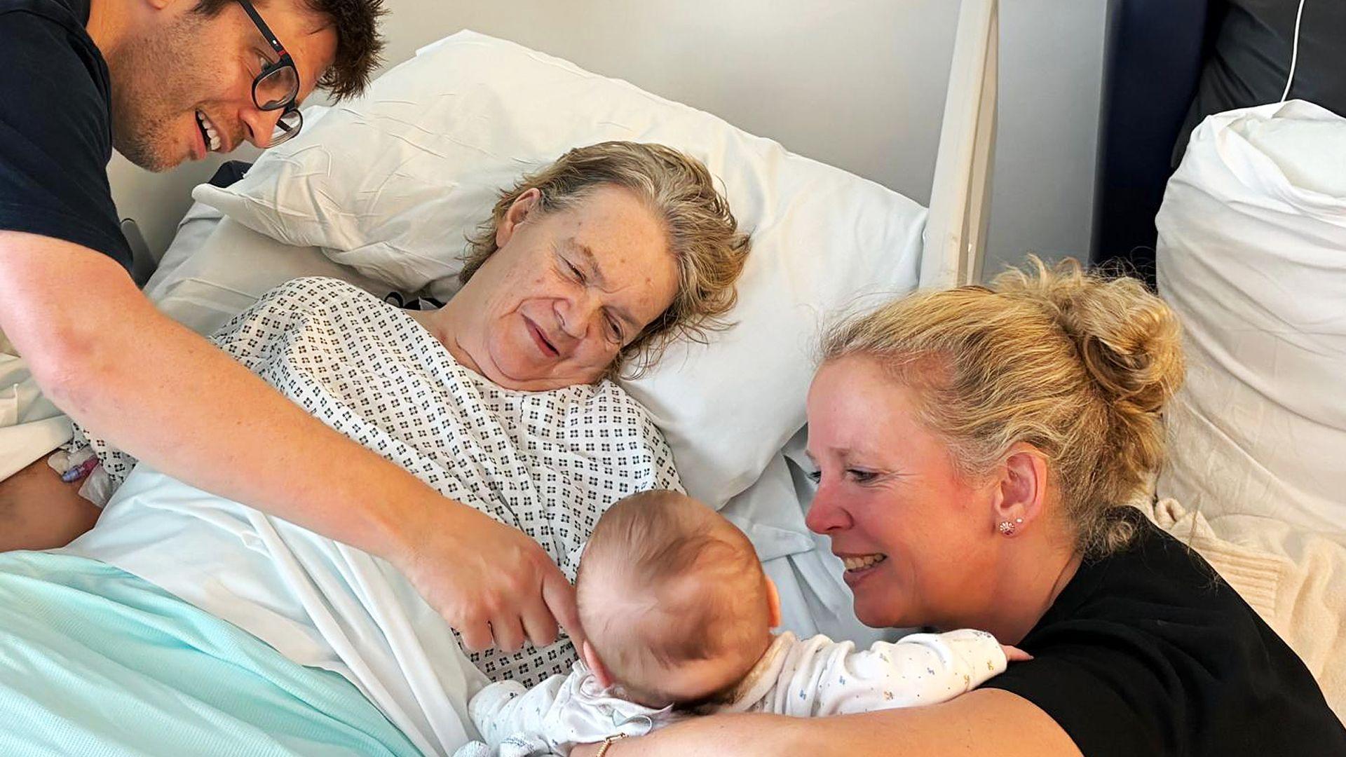 A woman in a hospital bed meeting her granddaughter. The baby's face can't be seen as the mother is holding the baby away from the camera, and a woman lying in the hospital bed is smiling at the baby. A man wearing glasses is leaning in and reaches his hand across the hospital bed to stroke the baby's cheek.