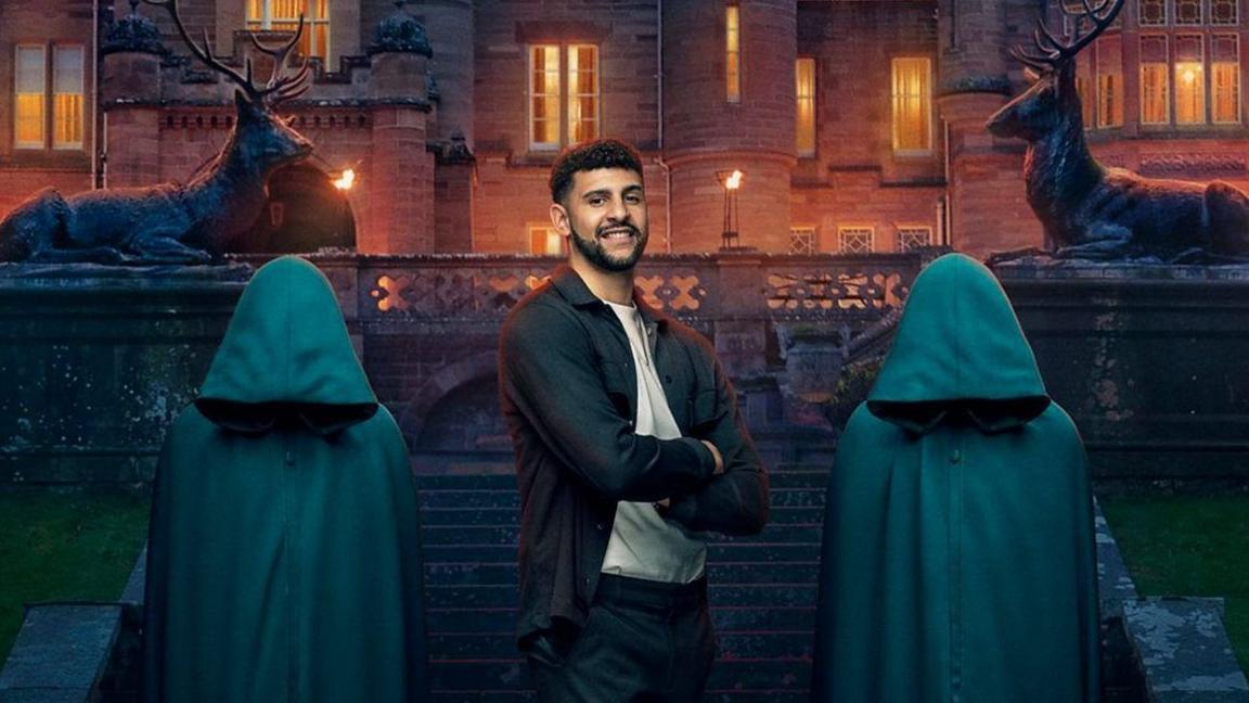 A man is smiling at the camera with his arms folded. He is standing in between two people wearing green cloaks and in front of a gothic castle