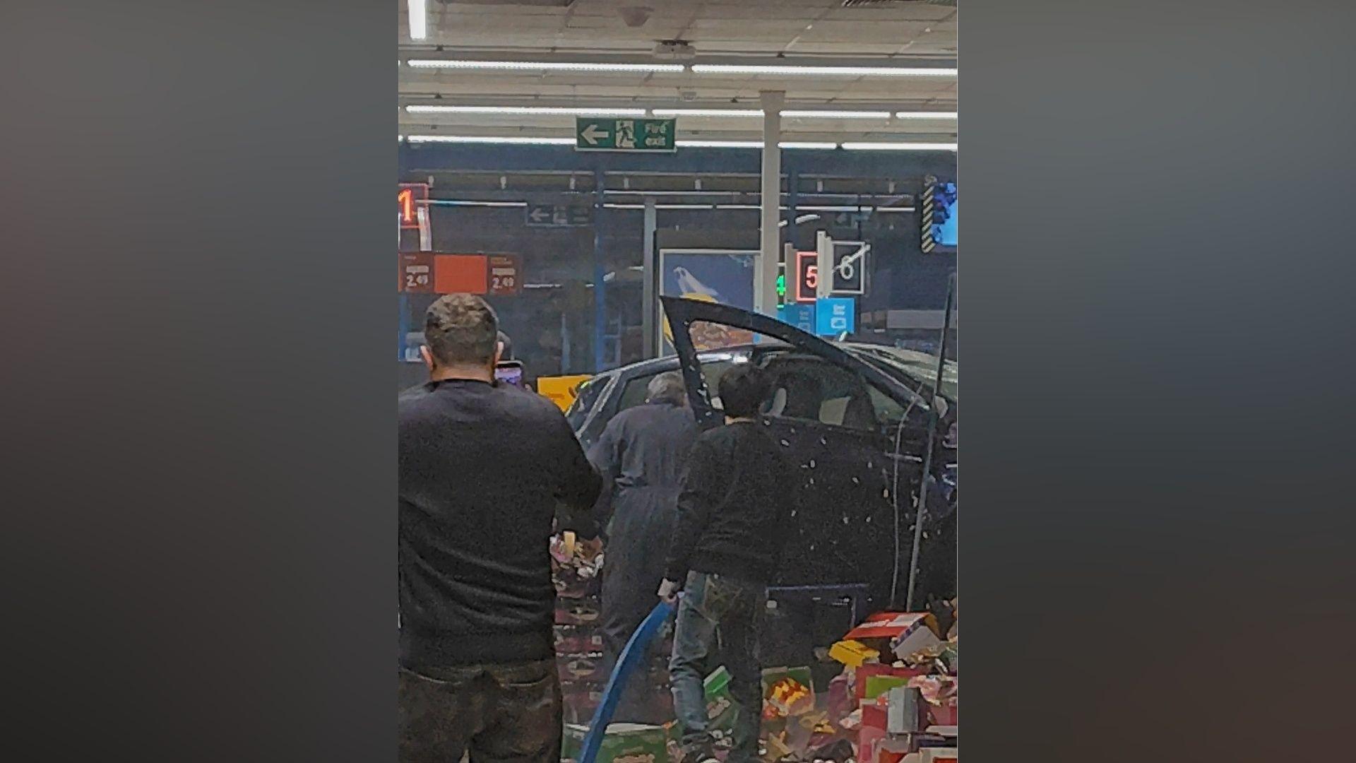 A car inside a supermarket with the backs of three people shown stood near it.