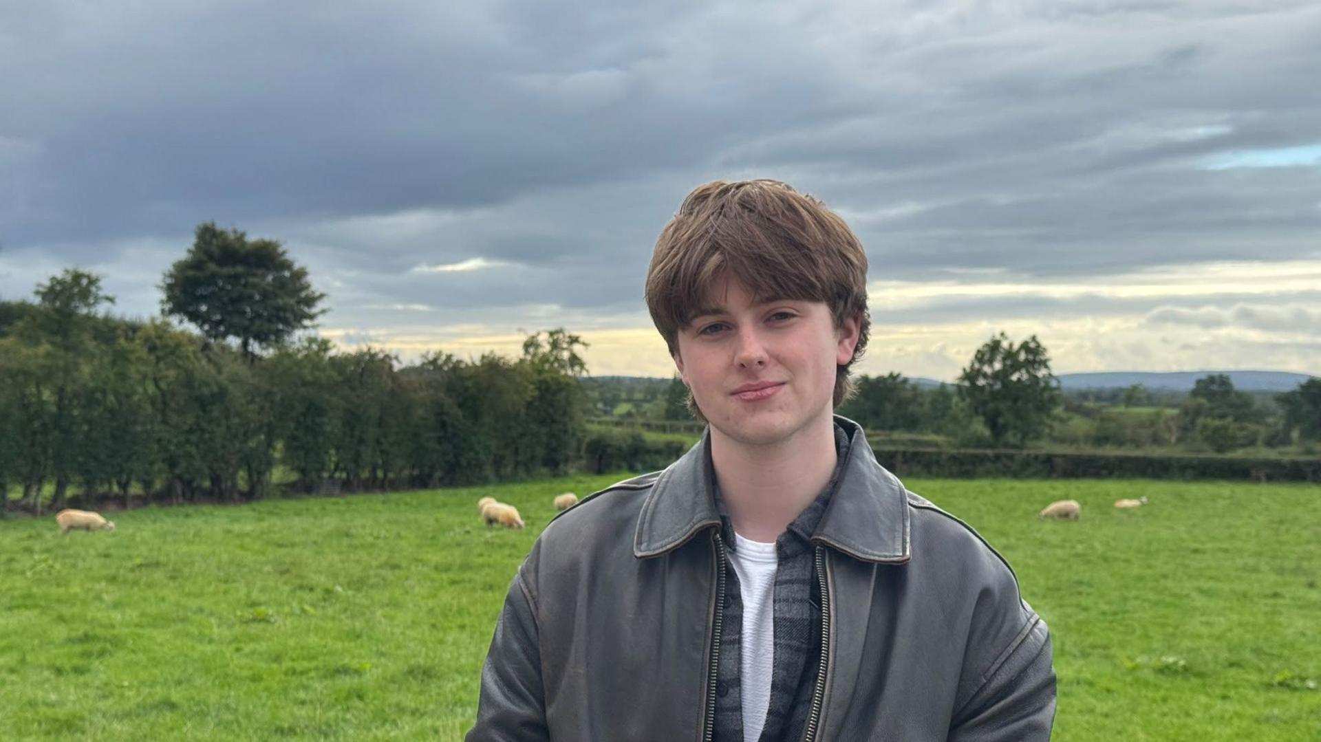 A young man in a leather jacket, with dark hair is in front of a field with sheep under a cloudy sky