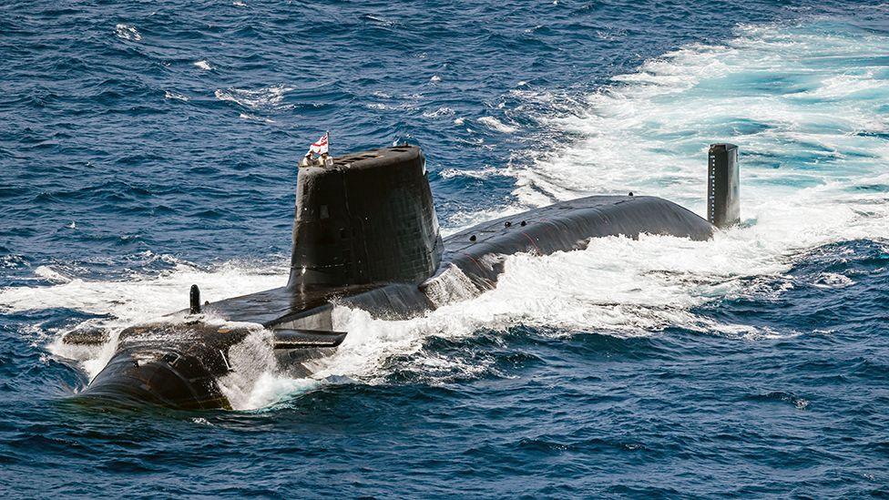 A black HMS Astute submarine, seen from above and at a slight distance so the whole vessel is visible and the size of it is unclear. A flag on the conning tower, two thirds of the way towards the front of the vessel, gives a hint at the scale of it. The submarine is surrounded by blue-black sea with churning white foam on its flanks and in its wake.