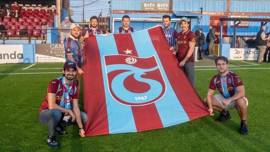 A group of Trabzonspor fans unfurl a flag at Drogheda's stadium, posing for the camera.