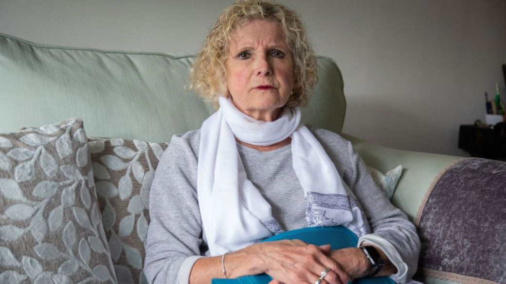 Sian Jones at home - a woman with a curly blonde bob, picture sitting of a grey sofa, holding a hot water bottle across her tummy.