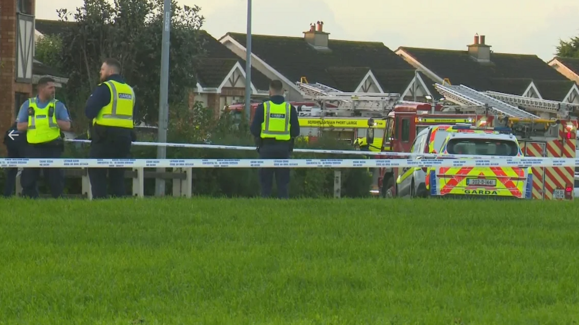 A number of gardaí (Irish police) stand guard at cordons in a housing estate in Waterford.  Fire engines are parked at the scene. The street is lined with large, semi-detached houses, some trees and there is a communal lawn in the foreground.