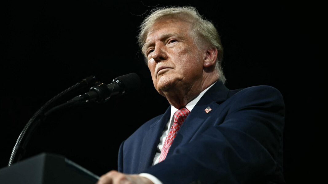 President Trump stands against a black background behing a black podium/ He is wearing a white shirt, blue suit coat with an American flag pin and a red print tie. He is not smiling and is gripping the edges of the podium. He is photographed from below the podium looking up.