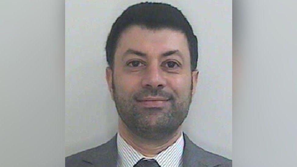 Image shows a Passport-style photo of Arif Patel, who has short dark hair, a short dark beard, and is wearing a grey suit with a grey and white striped shirt and grey tie.