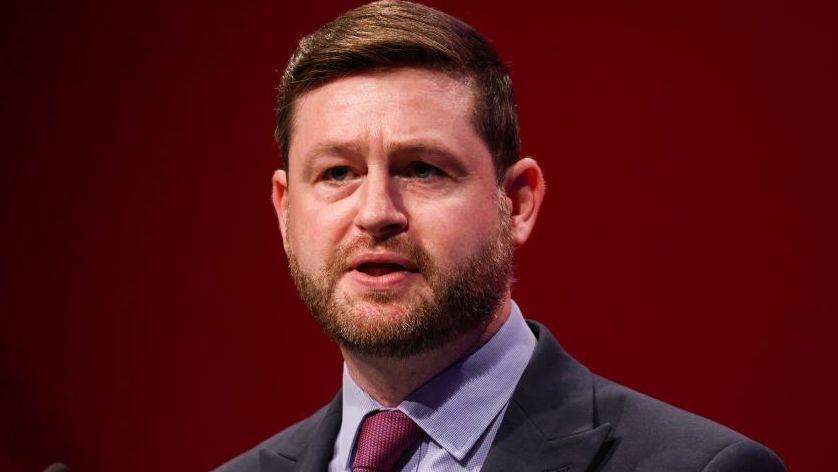 Head and shoulders shot of McMahon, who is speaking on stage with a burgundy backdrop. He is wearing a grey jacket over a blue shirt with a red tie.