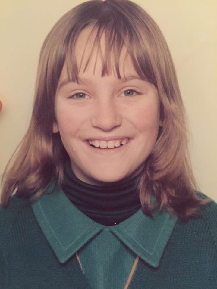 A studio-style portrait of Rachel as a child, shown from the shoulders up against a light plain background. Rachel has shoulder-length hair with a fringe and is looking directly at the camera, smiling. Rachel is wearing a dark roll-neck top under a teal-green collared jumper.