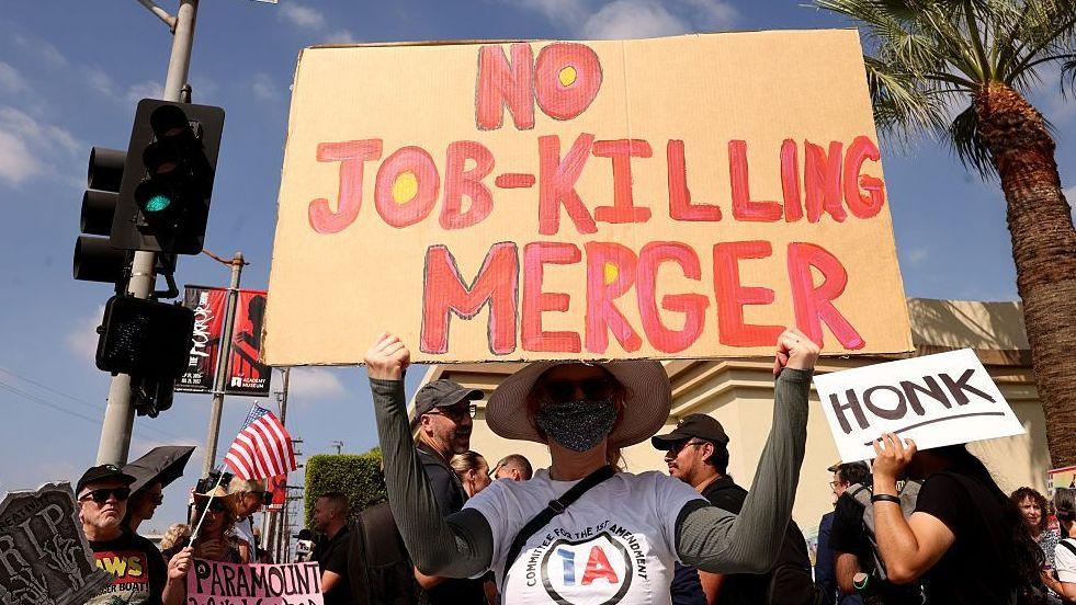 Protester holds a sign reading "No job-killing merger" in front of a crowd  outside Paramount Studios in Los Angeles in September, 2026.