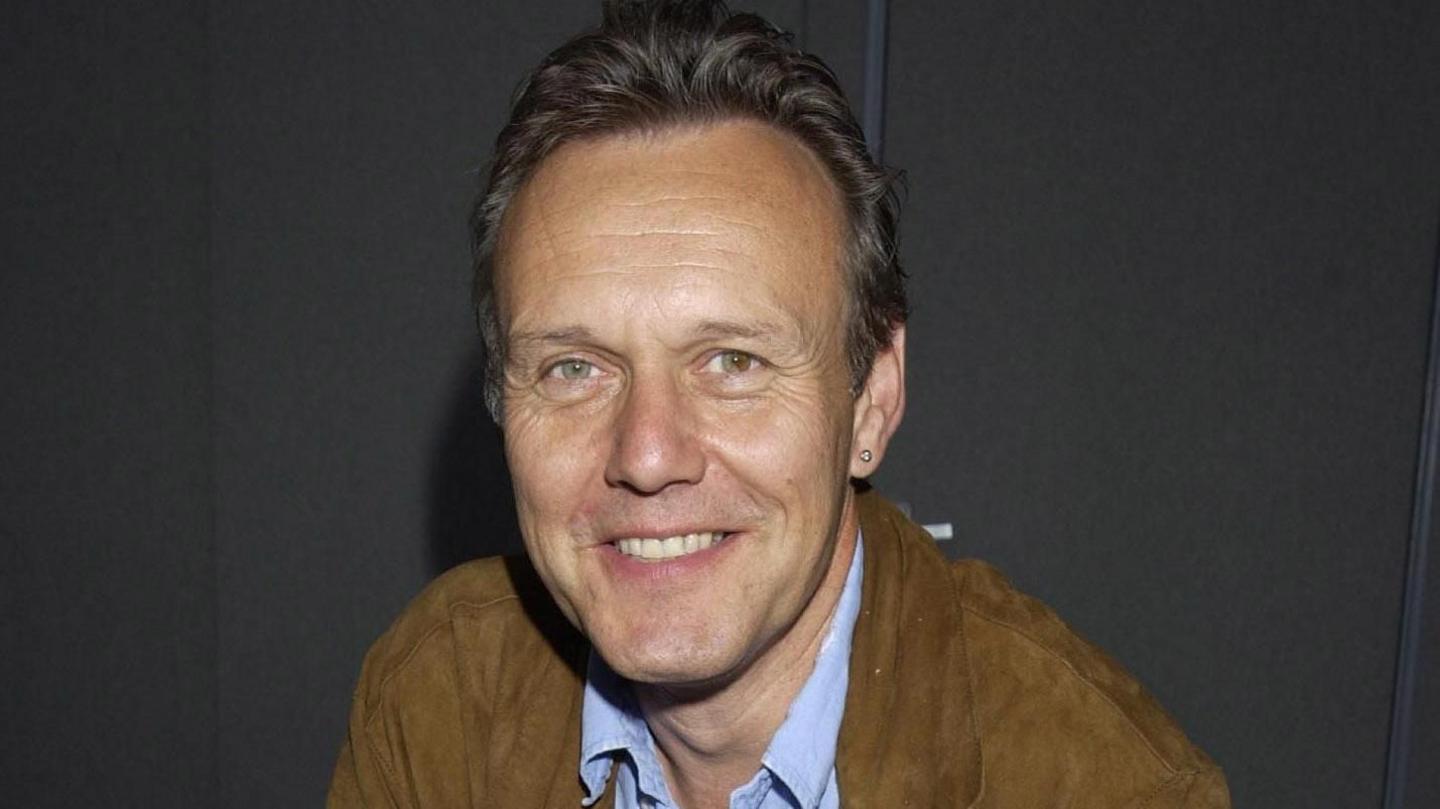 Anthony Head, a man in his early 60s with dark brown hair wearing a camel jacket and a blue shirt