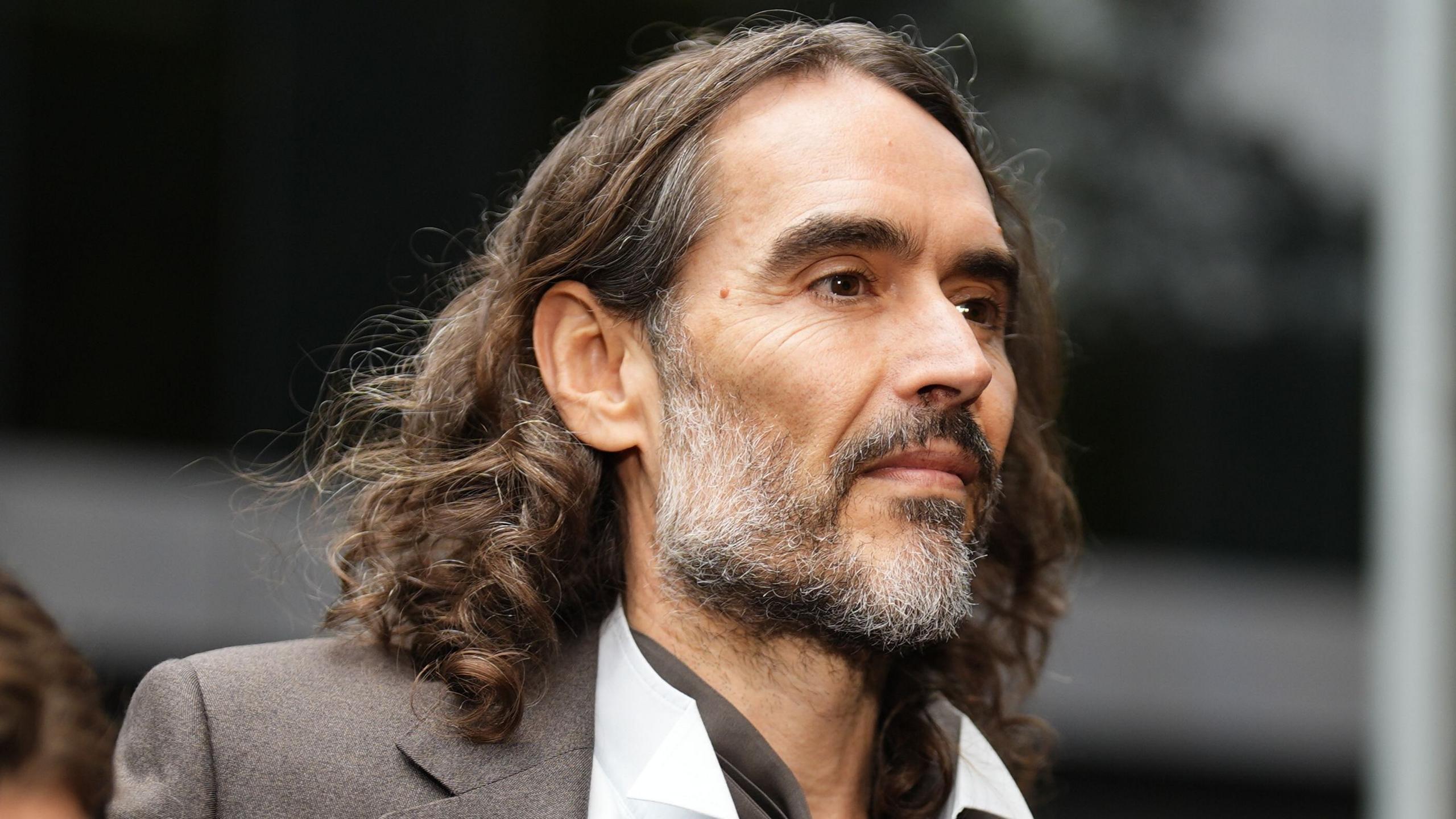 Russell Brand in a grey suit and open-necked white shirt arriving at court
