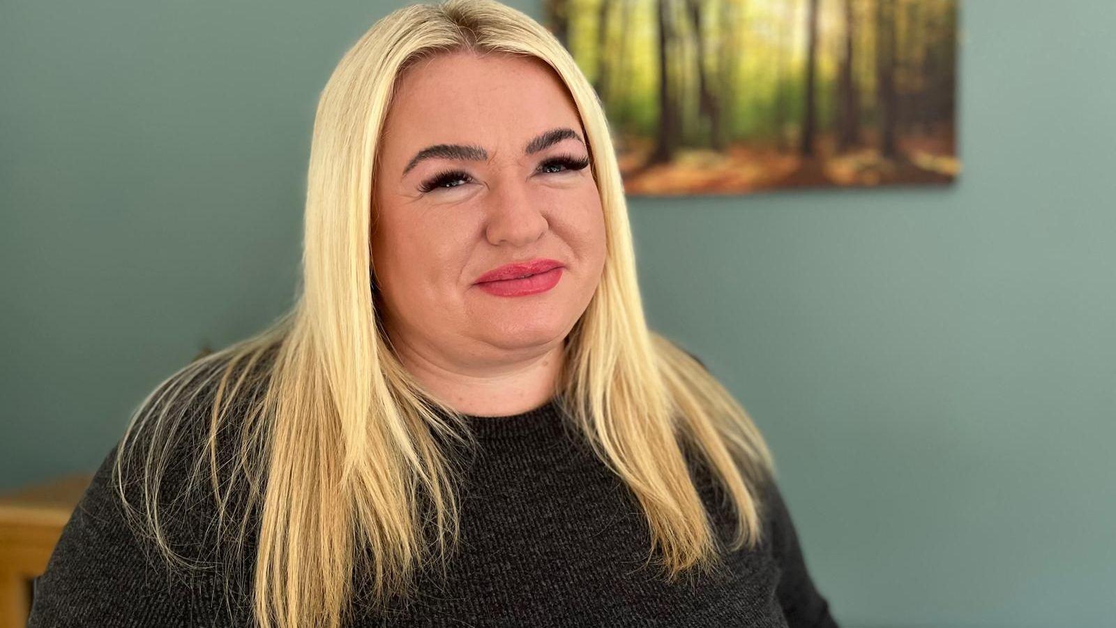 Jo has long blonde hair and is wearing a dark coloured dress. She is smiling at the camera. Behind her on the green wall is a picture of a wooded scene.