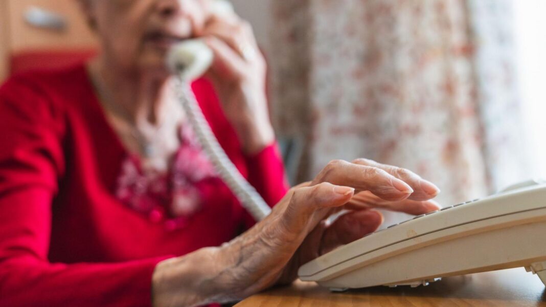 Older woman wearing a red cardigan dials a number on a push-button phone