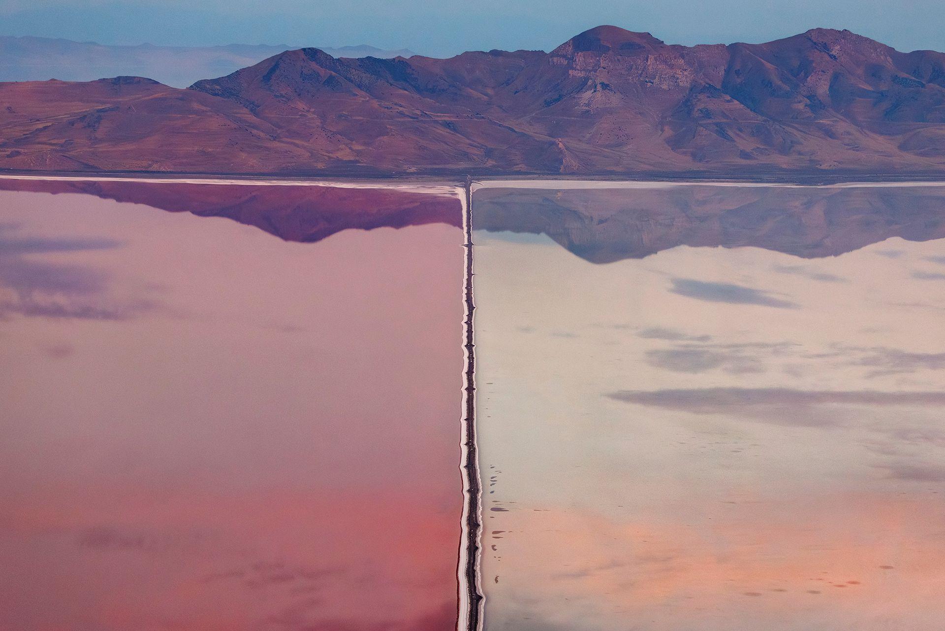 An aerial view of a divided lake stretches towards a rugged mountain range, with a narrow straight line running through the water from the foreground to the mountains. The water is pink on one side and pale blue-grey on the other, with the mountains reflected across the calm surface.