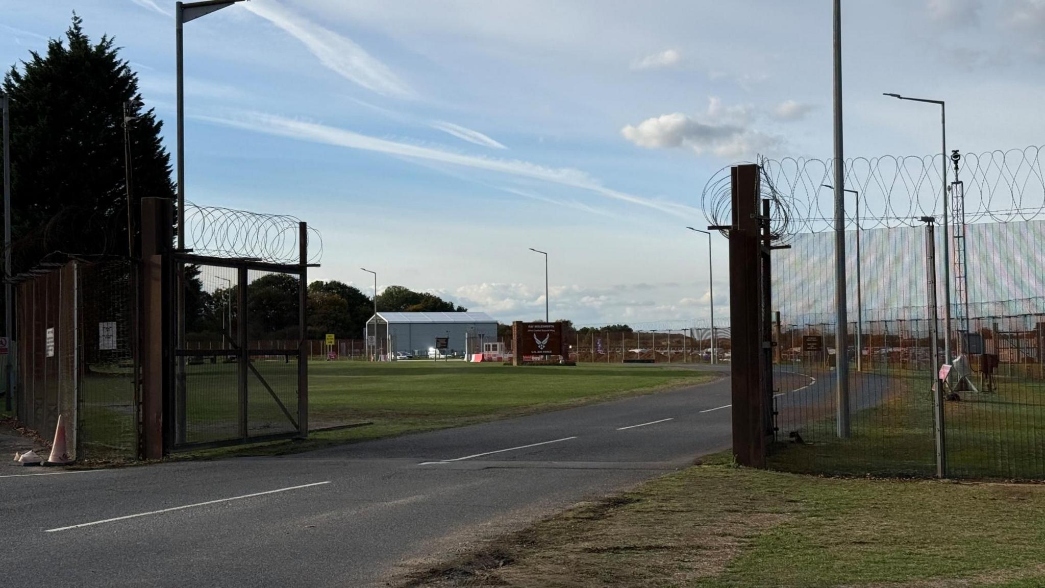 The entrance to an air base has open gates and barbed wire on the fencing. In the distance is a large building and more fencing.