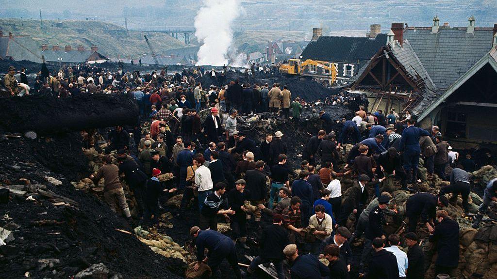 A colour photograph showing the damage to Pantglas Junior School, the ruined building is to the right of the frame. In the foreground is black coal waste and dozens of rescue workers