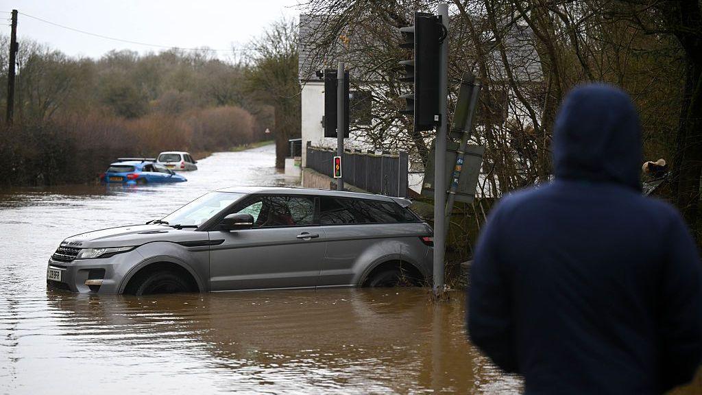 People look on at the cars stuck in the flooding in the hamlet of Weycroft, on January 27, 2026 in Axminster, England.
