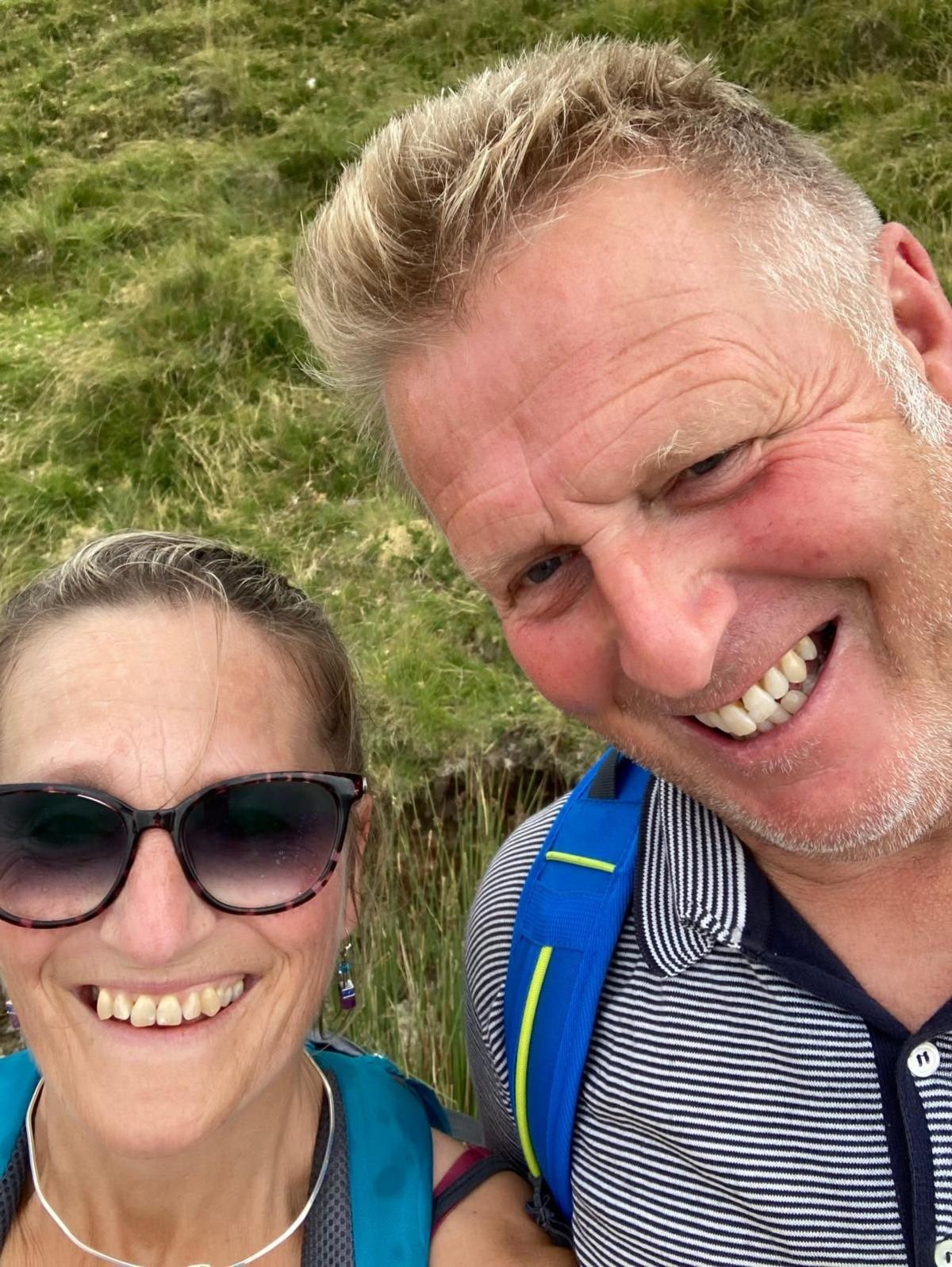 A close-up outdoor selfie showing Rachel on the left and Ian on the right. Both are standing on a grassy hillside and facing the camera. Rachel is wearing dark sunglasses, a blue backpack and a silver necklace. Ian is wearing a striped polo shirt and a blue backpack.