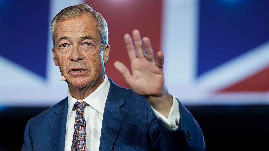 Nigel Farage holds his hand up while speaking on a stage with a Union flag in the background.