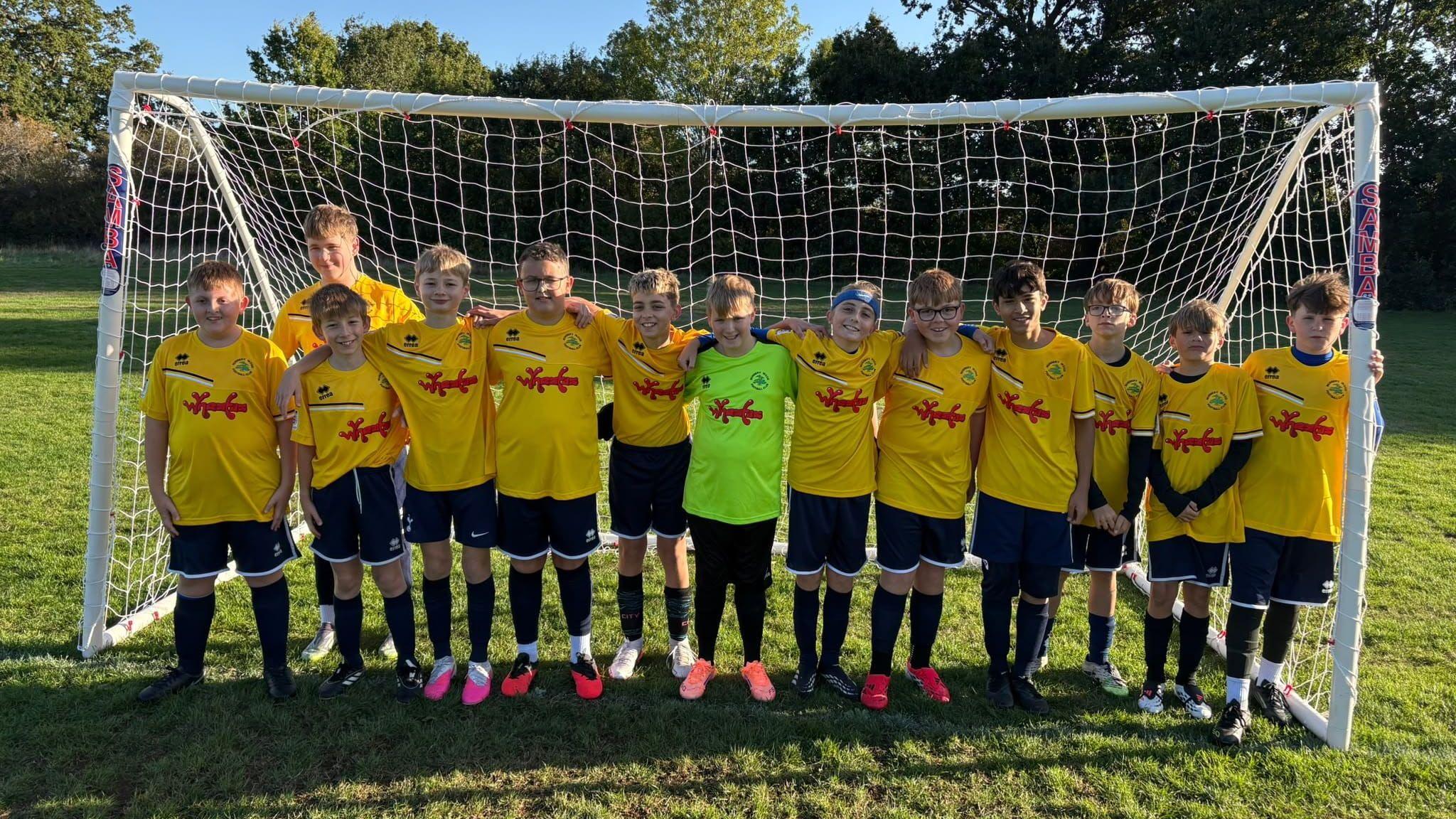 Thirteen young boys in yellow football shirts, black shorts and black socks line up in the goal mouth. They each have their arms around each other. The band's name is written in red writing across their shirts.