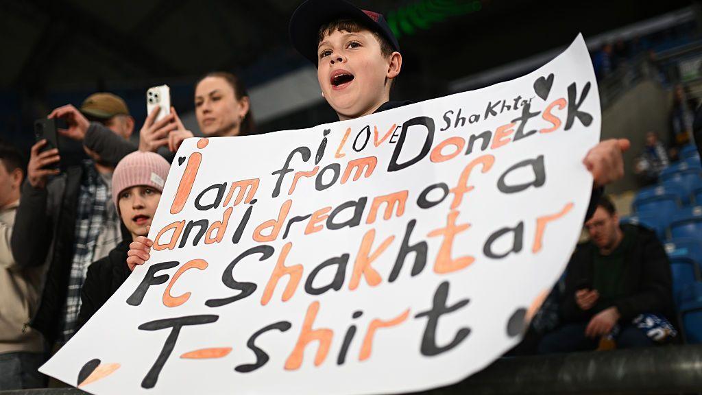 A young fan of Shakhtar Donetsk holds up a sign which reads "I love Shakhtar, I am from Donetsk and I dream of a FC Shakhtar Donetsk T-shirt" before a Conference League 2025-26 match against Lech Poznan