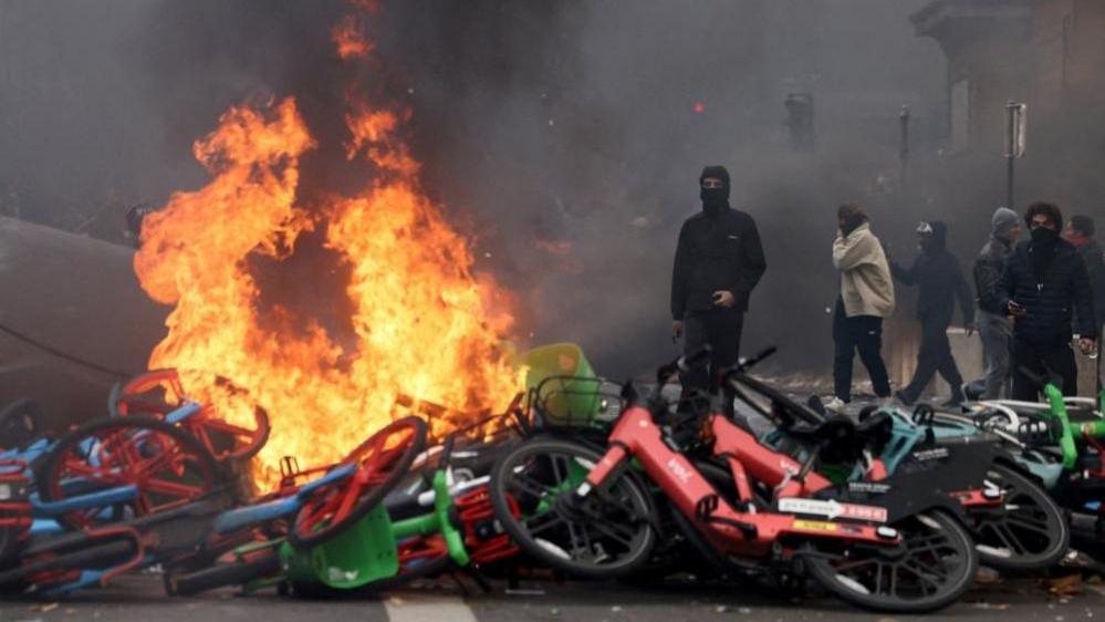 Masked protesters standing next to a pile of bikes, which has been set alight