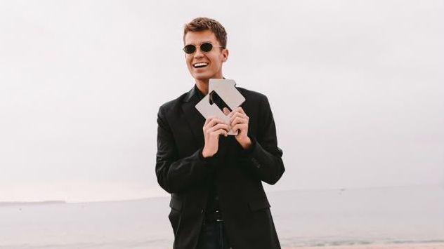 A man is wearing glasses and a black suit outfit, holding an award, on a sandy beach in front of the sea.