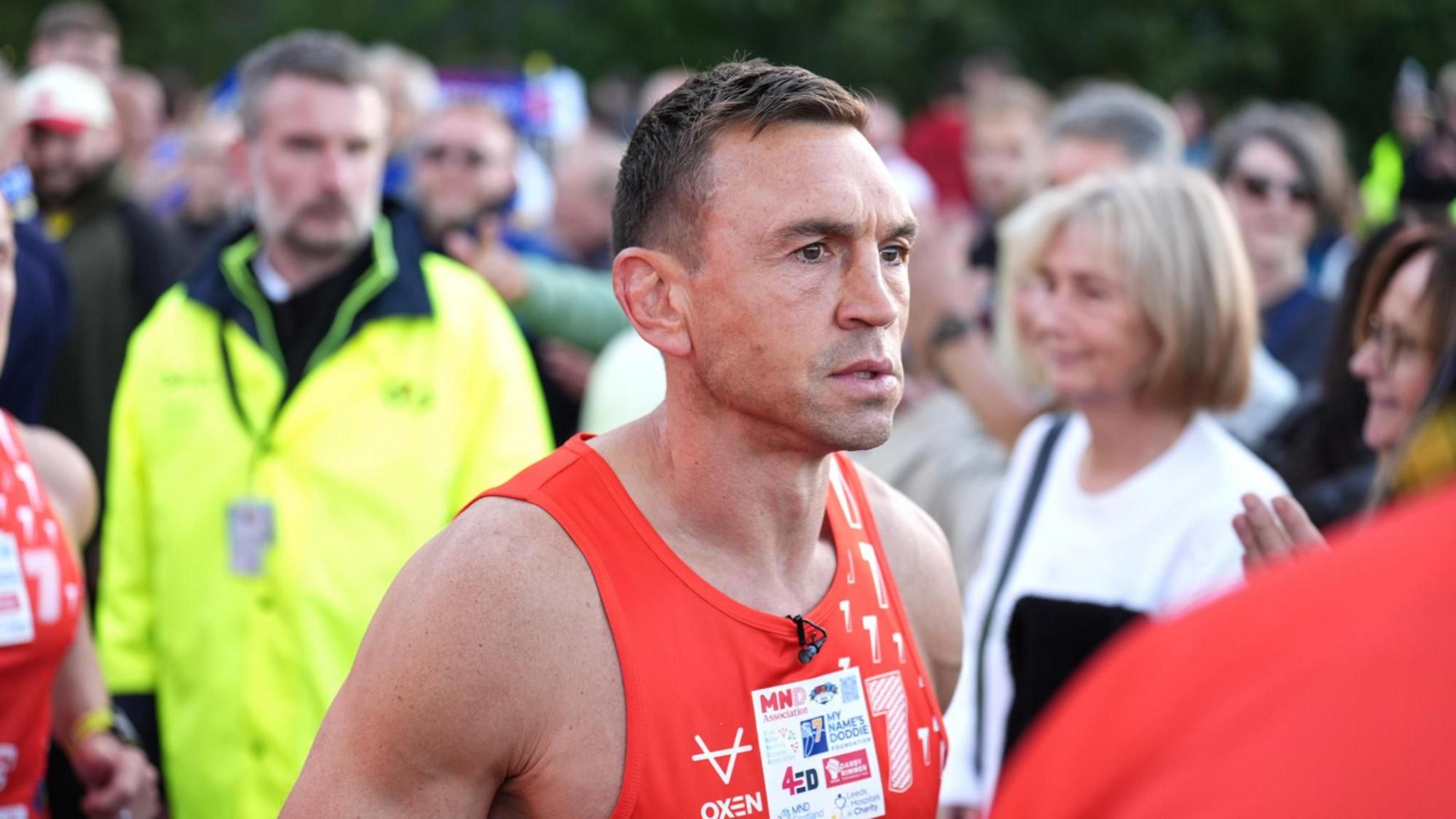 Sir Kevin Sinfield in a red running vest looks determined as a crowd gathers around him