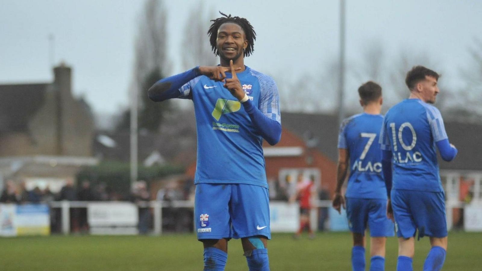 A man wearing a blue Rossington Main kit places one finger over the other to create a plus symbol while smiling. The move mimicks a goal celebration.