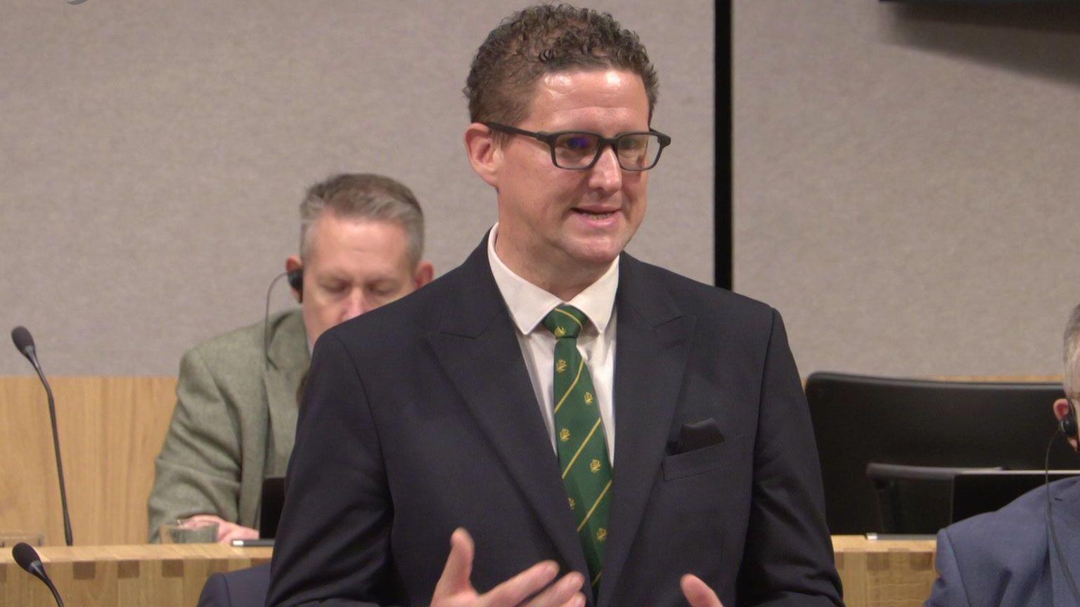 A man in a navy suit, green tie, and glasses, stands up and speaks inside a formal-looking room.