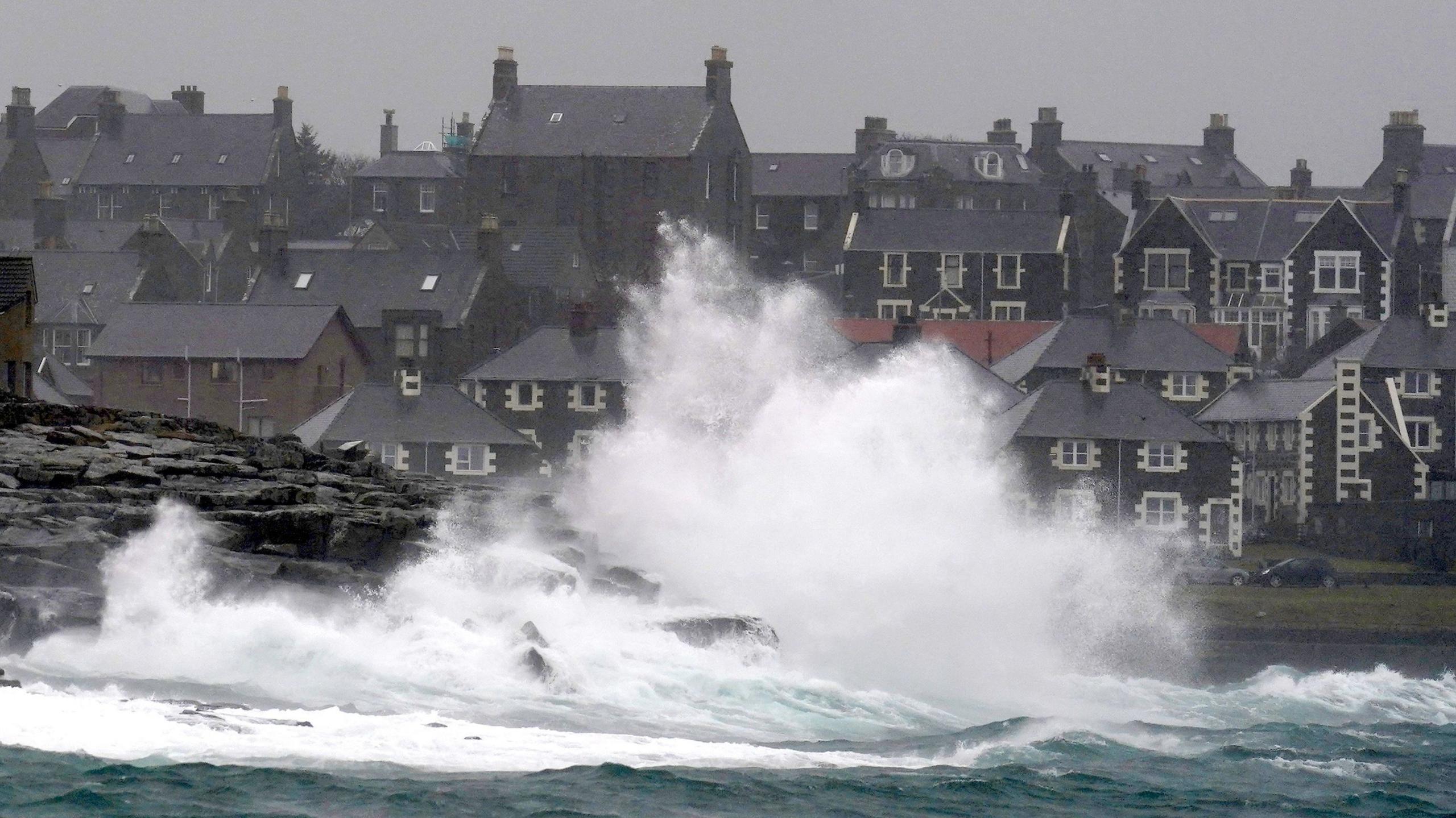 Waves crash over rocks and a seafront with houses in the background and grey sky overhead