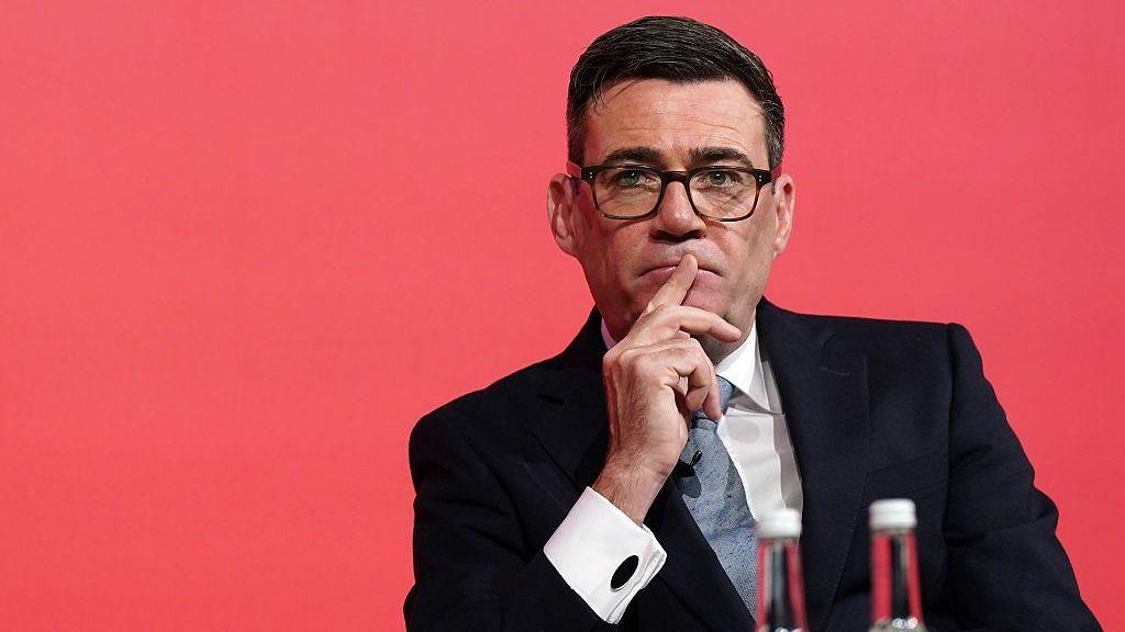 Andy Burnham looks pensive while sitting down with a red background.