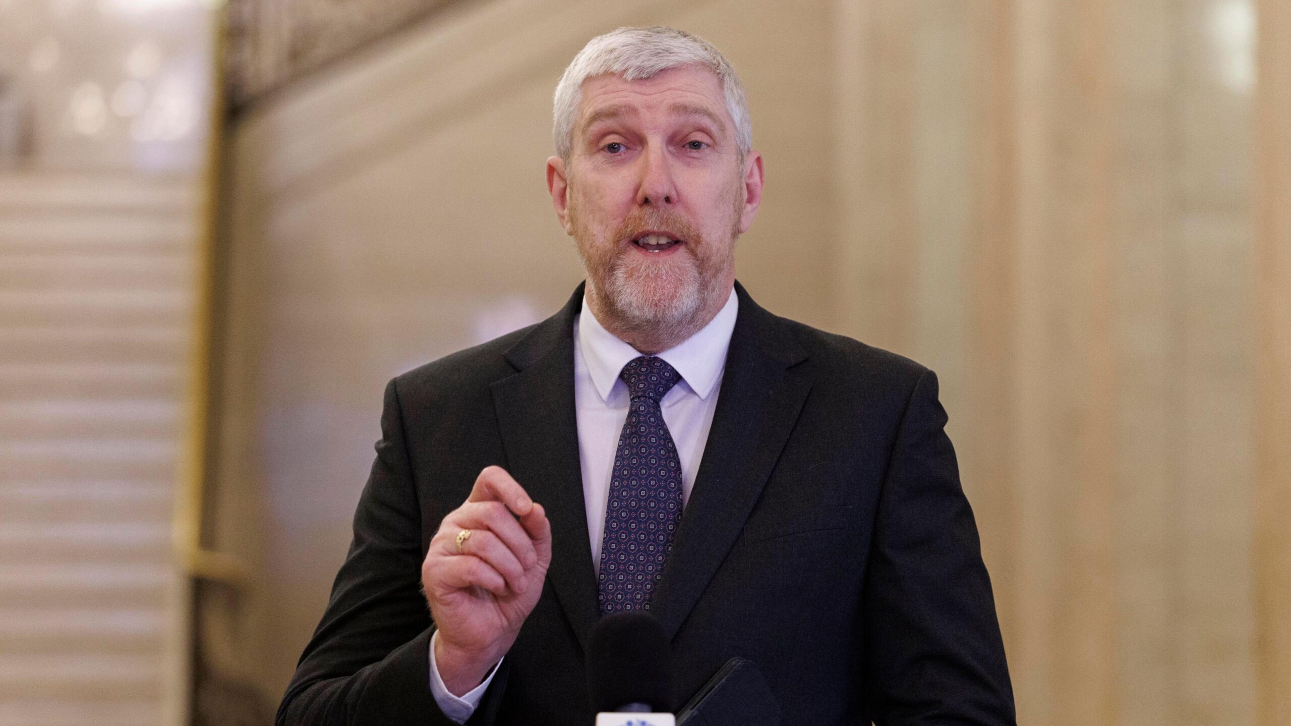 John O'Dowd speaking into a microphone in Stormont parliament buildings. He is wearing a black suit, navy patterned tie and a white shirt. He has grey hair.
