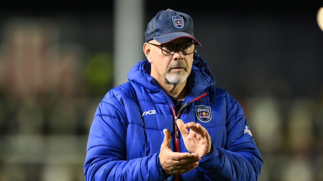 Dan McFarland clapping his hands together ahead of a Newcastle Red Bulls match. He is wearing a blue cap and a big blue puffa jacket with club logos on both