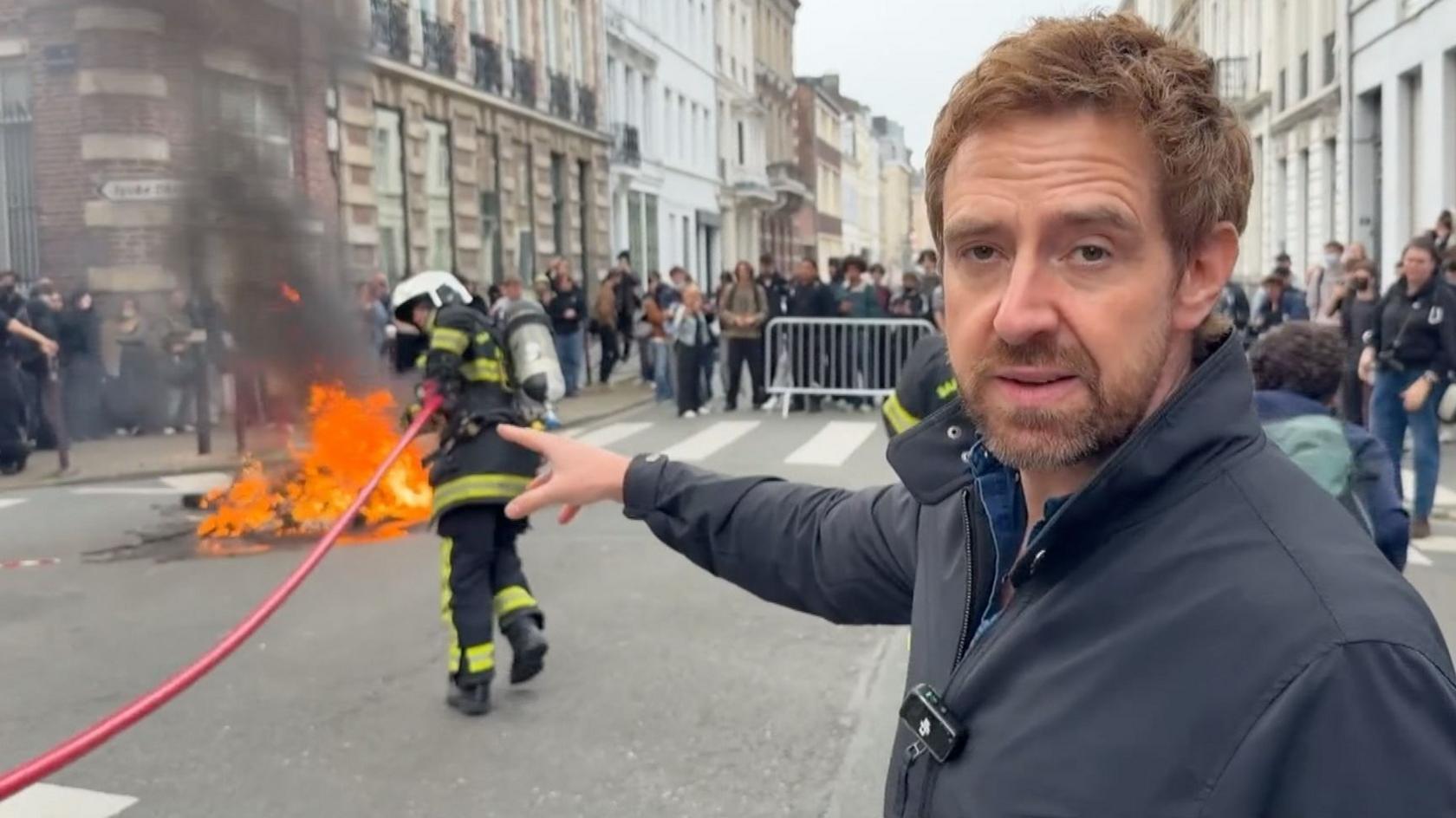 Nick Beake on a street in Lille with a burning bin behind him