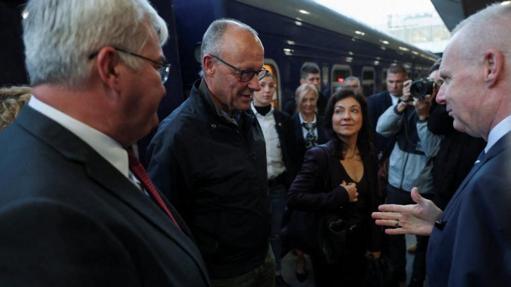 Friedrich Merz stands in the middle of a group of people in front of a train. They are looking at him and one man in the background can be seen taking a photo of him.