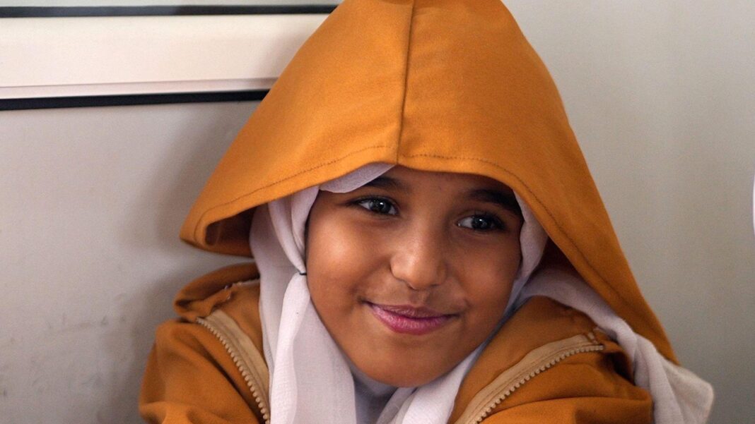 Children brave snipers and mortars to go to school near Yemen front line A young girl smiles shyly at the camera. She is wearing a dark yellow hooded coat with a cream headscarf.