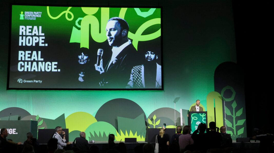 Greens face conference centre ban over Zionism motion A stage with green shapes on a green background and a large screen depicting two men and a woman with the message "real hope real change" to the left of them. A man in an olive suit stands at a lectern in front of it.