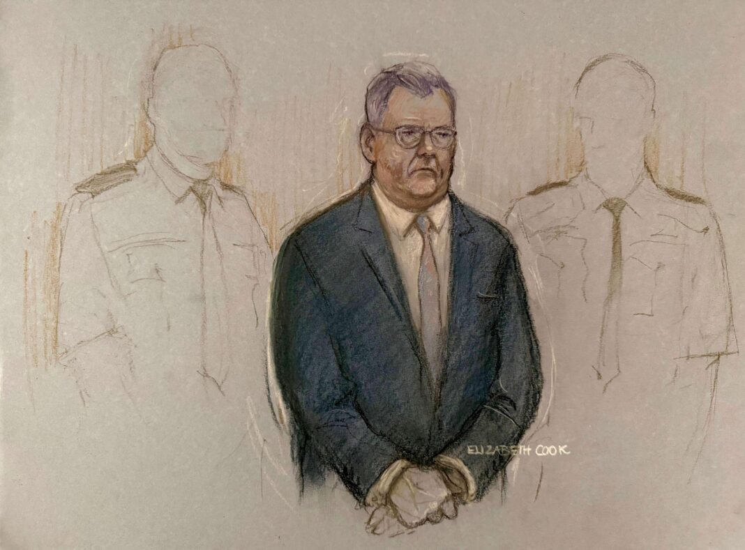 A court artist’s drawing. In the centre is a man wearing a navy suit and a tie. He is wearing a pair of glasses. The outlines of two faceless officers can be seen behind him.