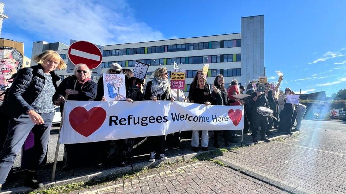 A group of protesters with various placards and a large banner saying Refugees Welcome Here
