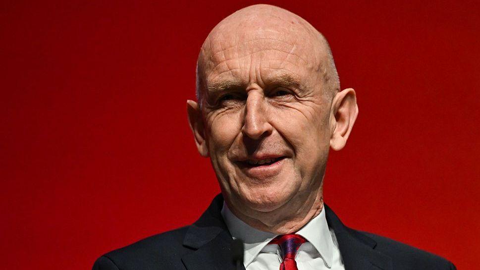 Chancellor John Healey wearing a dark suit, white shirt and red tie as he addresses the Labour Party conference