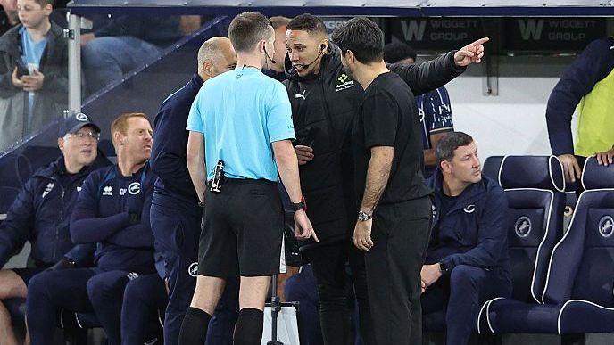 Referee Edward Duckworth in light blue shirt and dark shorts speaks to fourth official in black tracksuit, pointing with left arm