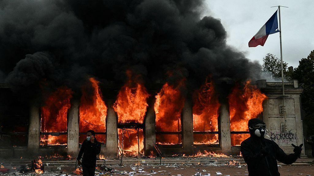 Two demonstrators, dressed all in black and in face coverings, stand outside the Lyon tourist office which is on fire behind them. A French flag is attached to the building.