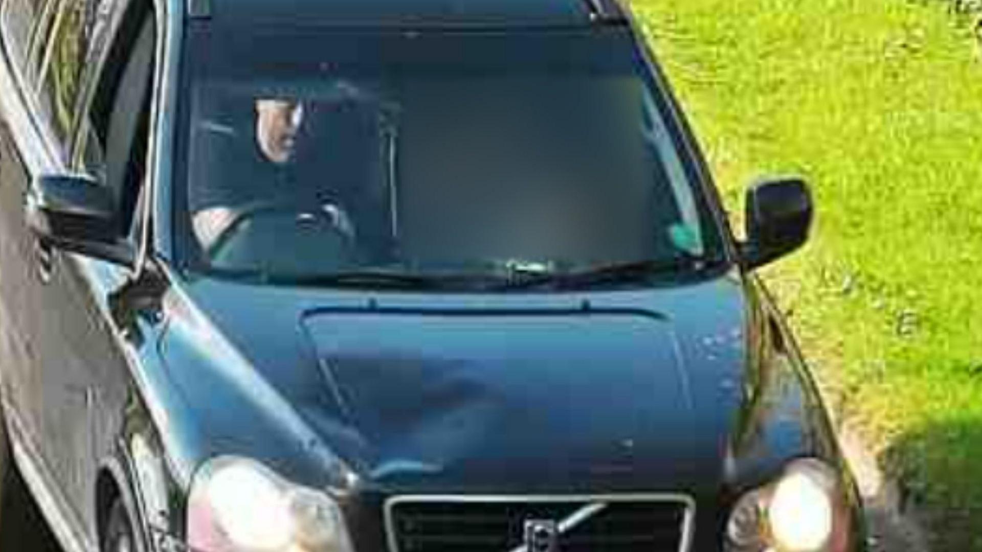 A CCTV image showing a black Volvo estate style car, with a visible dent in the bonnet and a cracked windscreen. A white man in his 30s, with dark hair and a black t-shirt is visible with his hand on the steering wheel. A woman in the passenger seat has been hidden by blurring.