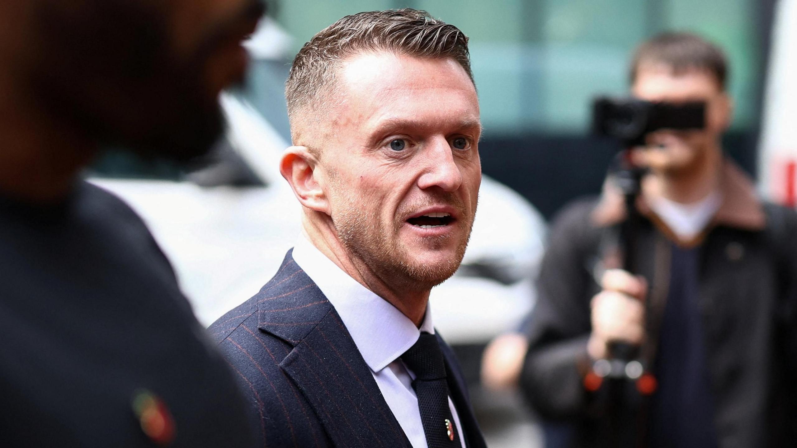 Tommy Robinson is walking towards the court, smartly dressed, surrounded by people, some filming him with cameras.