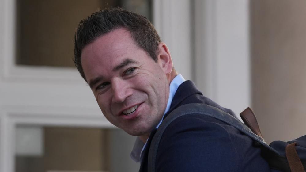 A man with short black hair wearing a suit jacket and blue shirt. He is looking over his shoulder as he walks into a building.