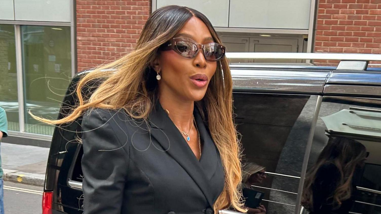 Naomi Campbell has long blonde and brown straight hair, brown sunglasses and a black blazer jacket. She is walking along, having just got out of a black car.