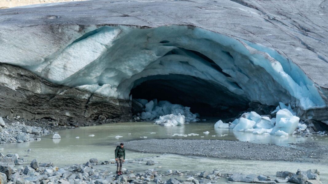 Swiss glaciers suffer ‘disastrous’ year of ice loss, threatening water supplies The front of a glacier, where a large channel - which looks a bit like a cave or an arch - has eaten away at the ice. A river of meltwater now meanders out of the front of the glacier. There are also chunks of ice on the ground having broken off the glacier. A man, dressed in mountaineering clothing, stands in front of the glacier for scale, which shows how big the channel or cave is.